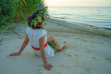 A girl of Slavic appearance with a wreath of wild flowers on her head sits on the shore of a lake....