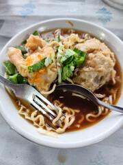 Bakso. A complete bowl of meatballs with yellow noodles, crispy fried dumplings and fresh green vegetables served with delicious, rich sauce on the table.