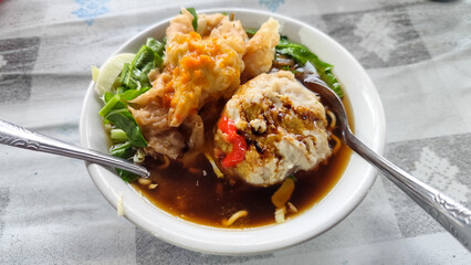Bakso. A complete bowl of meatballs with yellow noodles, crispy fried dumplings and fresh green vegetables served with delicious, rich sauce on the table.