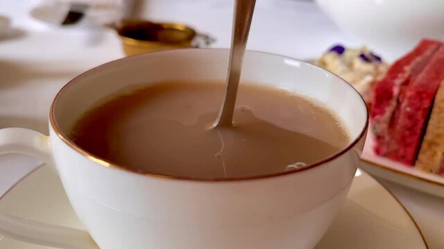 Freshly poured tea with milk swirled gently in fine porcelain, accompanied by dainty sandwiches and soft florals in a serene English afternoon tea moment, slow motion