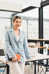 Young Asian businesswoman light blue blazer standing confidently in modern office holding notebook...