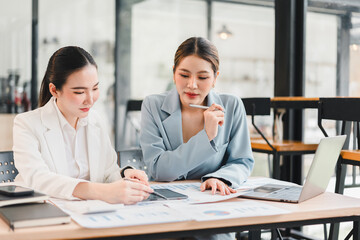 Two Asian businesswomen in formal suits discussing financial charts at modern office table, using laptop and tablet, focused and collaborative atmosphere