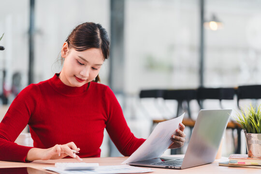 Young woman red sweater working at desk holding document using laptop modern office focused professional paperwork daytime natural light
