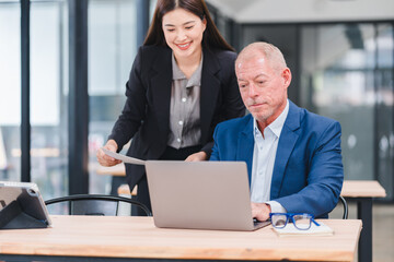 Young Asian businesswoman and senior Caucasian businessman working together in modern office, discussing documents and using laptop, teamwork and collaboration concept