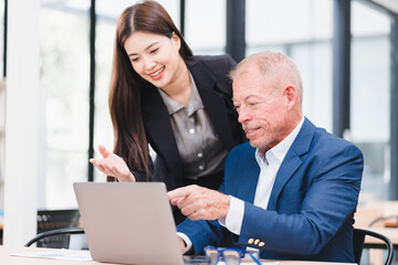 Young Asian businesswoman and senior Caucasian businessman working together in modern office, discussing documents and using laptop, teamwork and collaboration concept