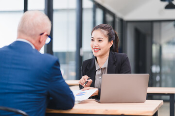 Young Asian businesswoman in black suit smiling and discussing work with senior male colleague in modern office, using laptop and documents, teamwork and professional collaboration concept