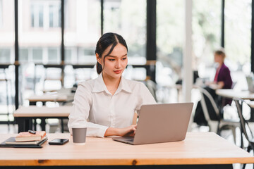 Obraz premium Young Asian woman working on laptop in modern office natural light professional atmosphere focused expression business casual technology productivity workspace