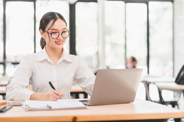 Young Asian woman, wearing glasses and white shirt, smiling while working on laptop and taking notes in modern bright office, professional and focused atmosphere