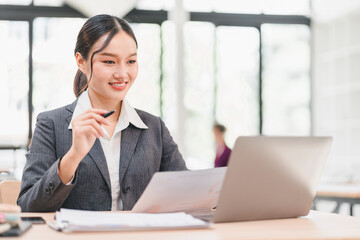 Fototapeta premium Confident businesswoman in gray suit working on laptop, holding document and pen, smiling in modern office with natural light, professional and focused atmosphere