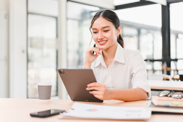 Fototapeta premium Young Asian woman in white shirt smiling while using digital tablet at modern office desk, with coffee cup, smartphone, and documents, showing productivity and positive mood