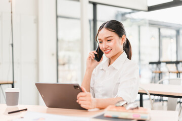 Young Asian woman business attire smiling working at modern office desk using tablet and talking on...