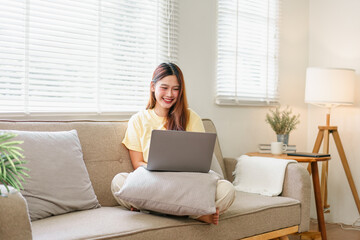 Young Asian woman sitting on beige sofa with laptop, smiling and working in bright living room with...