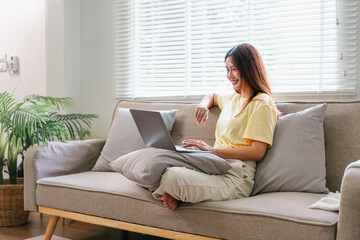 Young woman sitting on comfortable sofa with laptop, working from home in bright living room, casual and relaxed atmosphere, natural light from window, cozy interior