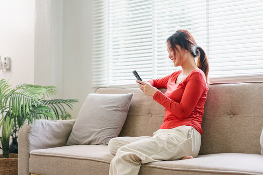 Young woman red shirt beige pants sitting on sofa using smartphone smiling relaxed bright living room natural light modern home interior casual atmosphere
