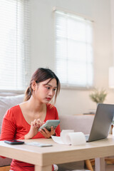 Obraz premium Young Asian woman in red shirt using calculator and laptop at home, focused on financial planning or budgeting, sitting at wooden table with paper receipt, natural daylight