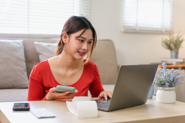 Young Asian woman in red shirt using calculator and laptop at home, managing finances with bills and receipts on wooden table, focused and positive atmosphere