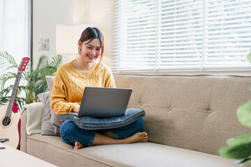 Young woman sitting cross legged on sofa with laptop, smiling and working in bright living room...