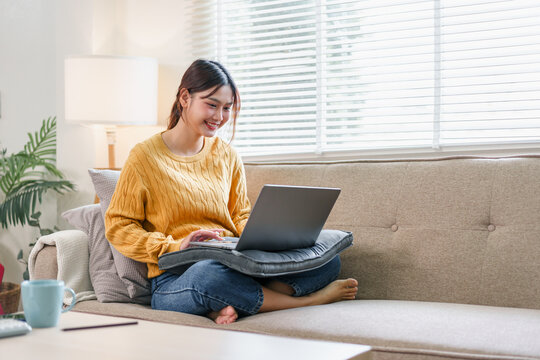 Young woman Asian sitting on sofa using laptop casual home interior natural light relaxed and happy mood remote work cozy living room modern lifestyle comfortable atmosphere - Powered by Adobe