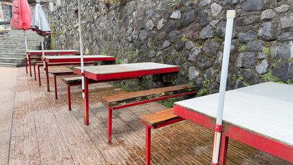 This image shows a row of outdoor picnic tables with red frames and wooden seats, set alongside a stone wall. Umbrellas provide shade, suggesting a dining area or rest stop