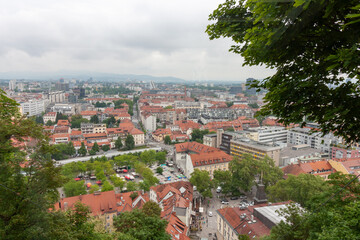 An expansive elevated view captures the cityscape of Ljubljana, on a cloudy day. Red-tiled rooftops...