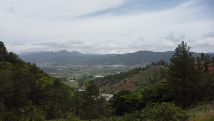 Panoramic view of a lush valley surrounded by forested mountains under a cloudy sky in Constanza, DR. Farmland patterns, green hills, and distant urban areas are visible in this scenic rural landscape