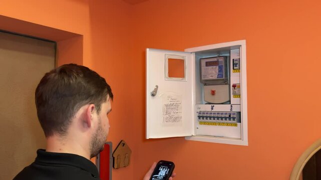 A man opens the door to an electricity meter and documents the readings by taking a photo with his smartphone. Scene captured in a home utility area.