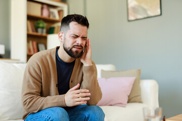 Caucasian Male Adult Showing Signs of Migraine Pain Seated on Living Room Sofa with Bookshelf...