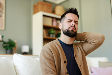 Young Man in Pain Holding Neck While Sitting in Cozy Living Room with Neutral Decor