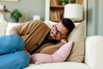 Caucasian Male Adult Experiencing Discomfort While Resting On White Sofa In Bright Home Interior...