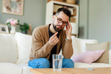 Bearded Man In Casual Attire Holding Medicine While Sitting Cross-Legged On White Sofa At Home