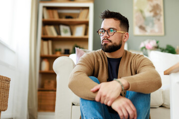 Thoughtful Young Caucasian Man in Glasses Sitting on the Floor of a Bright, Cozy Living Room