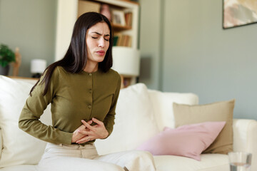 Young Woman in Green Sweater Holding Stomach in Pain While Sitting on a Comfortable Sofa