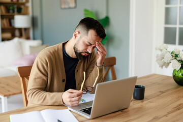 Stressed Caucasian Male Professional Rubbing Temple During Remote Work Session At Home Office Desk