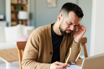 Young Caucasian Man in Pain Working from Home Holding Glasses in a Comfortable Indoor Environment