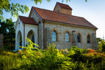 The Church of Saint Conrad of Parzham in Techirghiol, a charming stone Roman Catholic church with a reddish-brown tiled roof, nestled amidst lush vegetation under a bright blue sky.