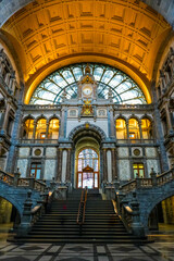 Fototapeta premium Interior view of Antwerp’s vaulted station hall, often called the “Railway Cathedral.” The wide marble staircase, arched portal and gilded clock sit beneath an iron-glass dome and golden ceiling.