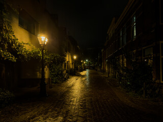 Night photo of a narrow alley in Leiden after rain. Gas-style lamps cast warm yellow light on wet...