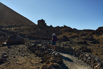Woman climbing a rugged volcanic trail toward the summit of Pico del Teide surrounded by dark lava...