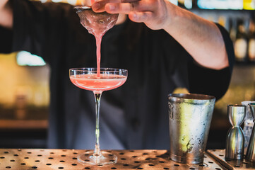 Bartender straining pink cocktail into glass at bar counter with metal shaker and jigger,...