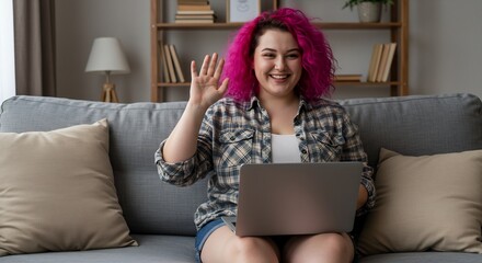Happy young woman with pink hair waving hello during a video call on her laptop while sitting on a comfy couch at home