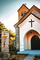 The Church of Saint Conrad of Parzham in Techirghiol, a charming stone Roman Catholic church with a reddish-brown tiled roof, nestled amidst lush vegetation under a bright blue sky.