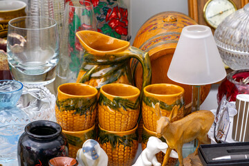 Corn cob mugs and jug surrounded by glassware and trinkets on a flea market stall.