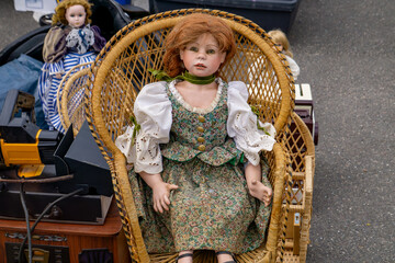 Vintage porcelain doll in a floral dress seated in a wicker chair, displayed among toys and...
