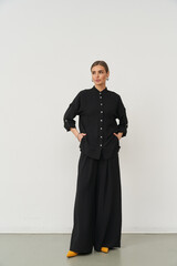 A woman with a neat bun wears a black button-up shirt and wide-legged black trousers, standing...