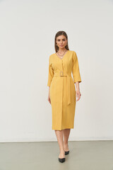 Yellow belted knee-length dress worn by a woman with long brown hair. She wears black shoes and...