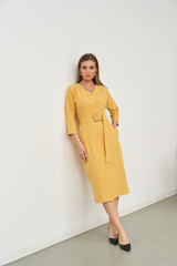 Woman in a yellow long-sleeved dress with a belt, standing against a plain backdrop. She wears...