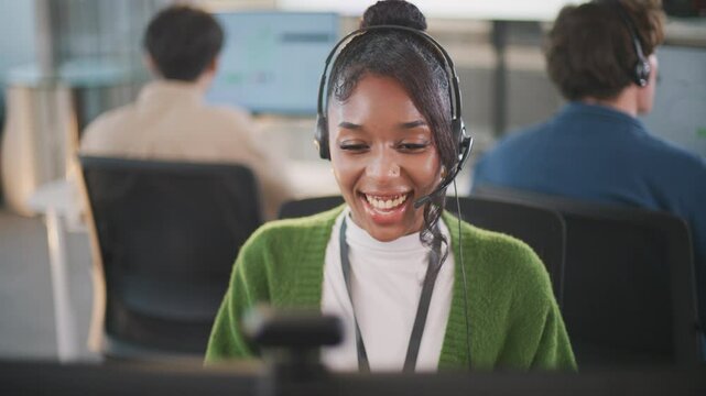 Confident African American female call center agent communicates with clients using headset and computer. She offers professional tech support at a modern office desk, smiling diverse office
