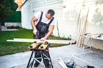 Mature male carpenter using electric drill on a wooden plank outdoors