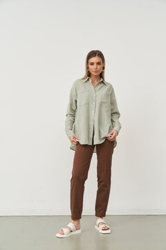 Neutral expression, woman stands confidently wearing a light gray button-up shirt and brown trousers with white sandals against a plain backdrop. She holds the shirt lightly with both hands