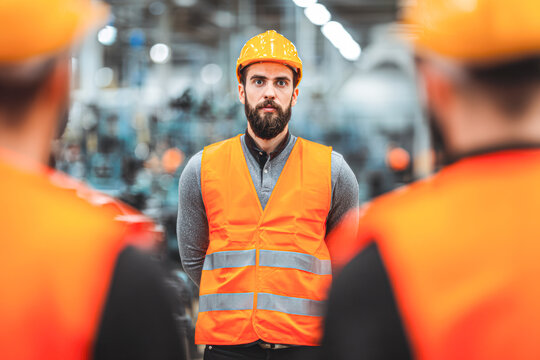 A man wearing a yellow hard hat and orange reflective vest stands confidently in a factory setting, facing forward with two blurred figures in safety gear in the foreground.